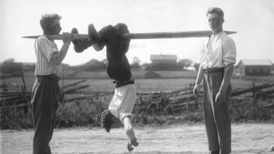 En man leker "flå katt", där han hänger upp och ner på en stång och ska ta sig framåt med att "hoppa" med benen. Bilden är tagen 1930 i Jomala, Björsby, Åland.