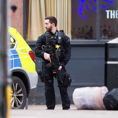 En polis med stort vapen står på en gata i Streatham, London, och ser mot vänster mot en polisbil. 