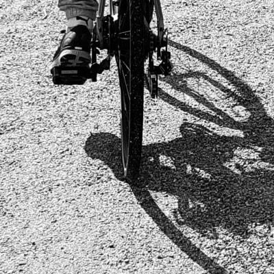 En fot som trampar en cykel över grus och cykelns skugga är enorm på grusplanen.