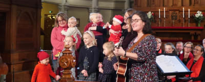 Musiklekgrupp med julklädda barn sjunger framme vid altaret i Johannes kyrka i Helsingfors. Delvis är de i mammors famn och leds av en kvinnlig gitarrist.