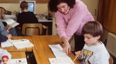 Opettaja ohjaamassa oppilaita harjoitustehtävien tekemisessä 1990-luvulla. Taustalla oppilaita tietokoneen ääressä