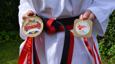 En kvinna i taekwondodräkt håller i två medaljer framför ett svartrött bälte.