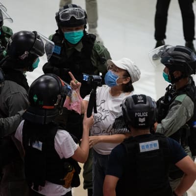 Mellakkapoliisit piirittävät ostoskeskuksessa naista, jolla on kasvosuojus ja joka heristää sormeaan poliiseille.