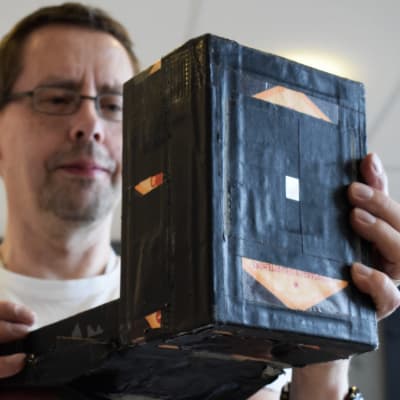 Hans G Hästbacka håller upp en nålhålskamera, en svarttejpad låda, stor som en större bok, framför sig.
