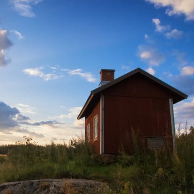 En röd stuga på en klippa med gräs i solnedgång.