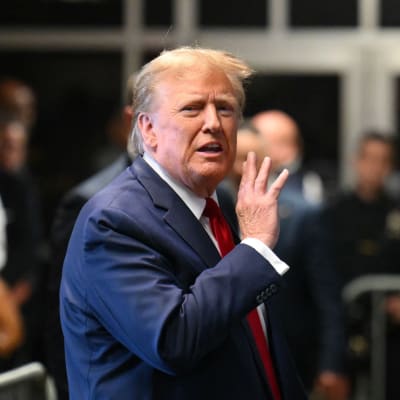 Donald Trump talar inför en publik i mörkblå kostym, och lyfter på sin högra hand mot munnen.