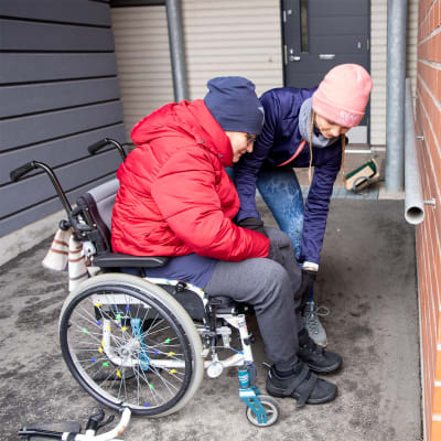 Avustaja auttaa Teija Kangasvieriä nousemaan pyörätuolistaan.