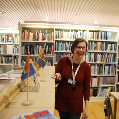 Kirjastonhoitaja Tiina Mäntylä seisaallaan kirjastossa, saamenlippuja pöydällä.