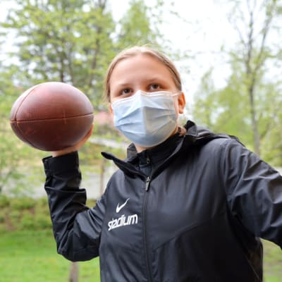 Julia Helppolainen i munskydd och svart jacka med en amerikansk fotboll i handen. 