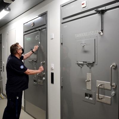 en man i skjorta, munskydd och glasögon låser upp flera lås till en fängelsecell, på bilden syns tre gråa dörrar till fängelseceller
