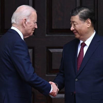 Yhdysvaltain presidentti Joe Biden ja Kiinan presidentti Xi Jinping kättelevät ennen tapaamista, taustalla tumma ovi.