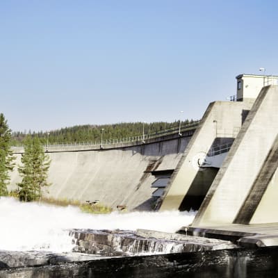 En femton meter hög damm i en fors, till vänster en skog, och till höger ett vattenkraftverk inbyggt i dammen med vatten forsande ut ur det.