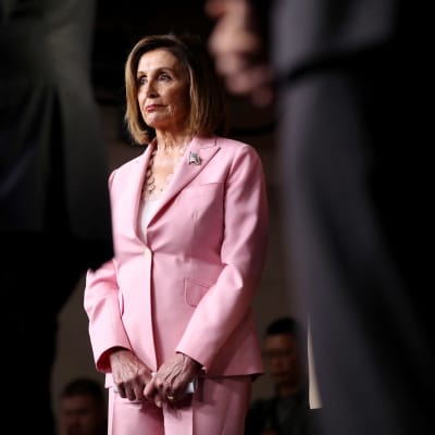 Nancy Pelosi, talman i USA:s representanthusets, iklädd rosa kostym står mellan svartklädda män. 