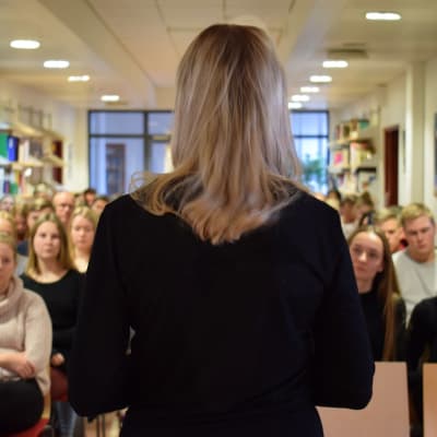 Sandra bergqvist med ryggen till kameran står framför en gymnasieklass och talar.