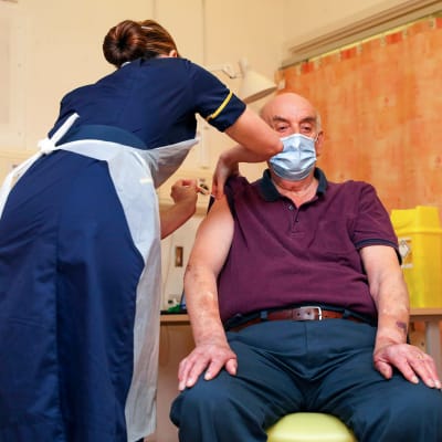 Bild på man i lila tskjorta. Mannen får vaccin av sjukskötare.