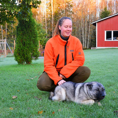 Kvinna i jaktkläder med hund framför sig på gräsmatta.