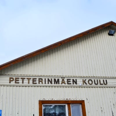 Närbild av en gammal träbyggnad, beige färg, det står Petterinmäen koulu på väggen.