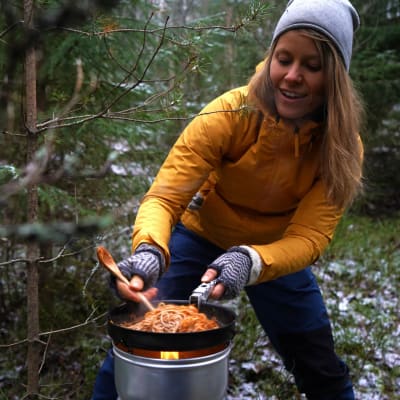 En kvinna med gul jacka och grå mössa lagar mat på ett stormkök i skogen.