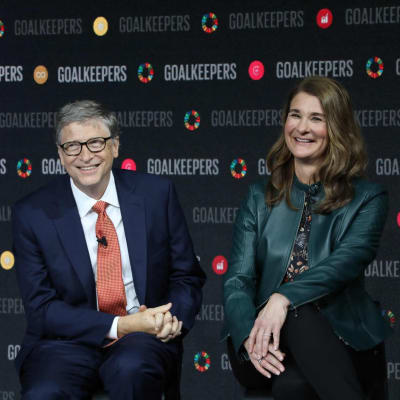 Leende Bill och Melinda Gates poserar sittande framför reklamvägg som marknadsför en av deras evenemang.