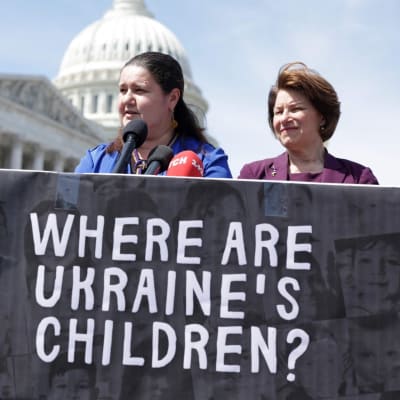 Ukrainan Yhdysvaltain suurlähettiläs Oksana Markarova seisoo kahden yhdysvaltalaispoliitikon välissä ja pitelee mustaa banderollia.