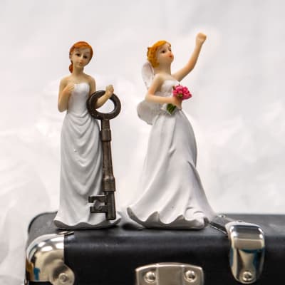 Tårtdekoration bestående av ett par kvinnor iklädda brudklänningar som står ovanpå en portfölj. Den ena har en nyckel i handen och den andra en blombukett.