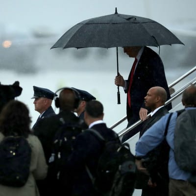 Presidentti Donald Trump astuu alas lentokoneesta sateenvarjon alla. Ympärillä on lehdistöä ja turvamiehiä.