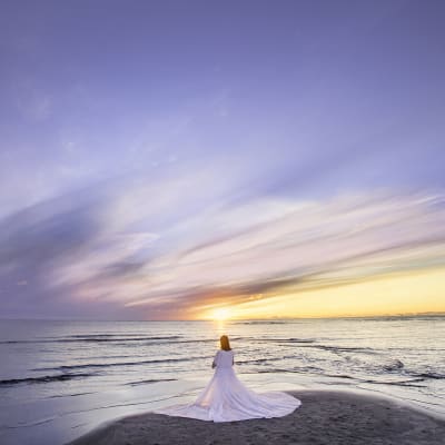 Valkoisiin pukeutunut nainen seisoo meren rannalla auringonlaskiessa