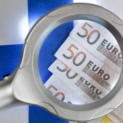 Bildsättning. Förstoringsglas över 50-eurossedlar, Finlands flagga i bakgrunden.