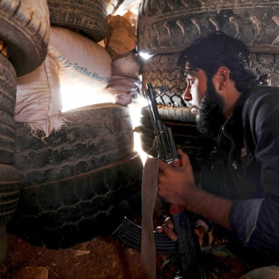 Syyrian kapinallistaistelija  tarkkailee etulinjaa autonrenkaista tehdystä suojasta.