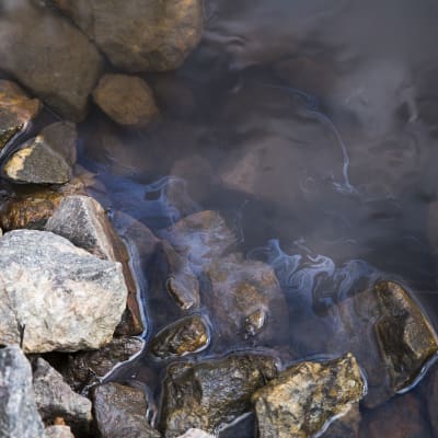 Bild på en bäck och stenar, på vattenytan syns skimrande olja.