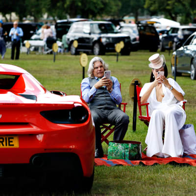 Harmaahiuksinen mies ja valkoisiin pukeutunut nainen istuvat punaisilla ohjajantuolilla ja näppäilevät puhelimiaan. Heidän vierellä vasemmalla on punainen Ferrari.