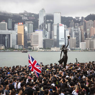 Valtava ihmisjoukko. Keskellä patsas. Taustalla Hongkongin pilvenpiirtäjiä.