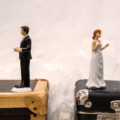 Ett bröllopspar (två figurer, kakdekorationer) står på kappsäckar och tittar bort från varandra.