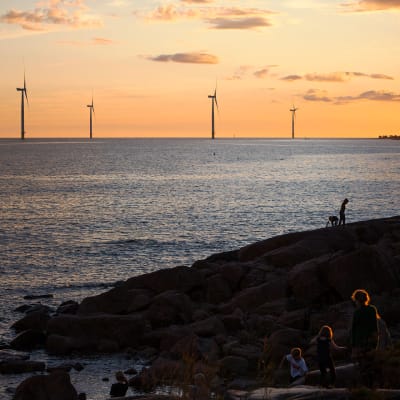 vindmöllor syns i en solnedgångshorisont vid havet. Några människor går på några klippor vid havet.