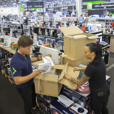 Två försäljare i en elektronikaffär packar upp kartonglådor mitt inne i affären. Hyllor med hemelektronik syns i bakgrunden.