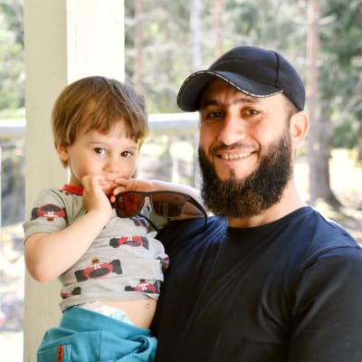 Adam Aki i grå t-skjorta med bilar på i famnen på Abdul Aki i svart t-skjorta och keps. 