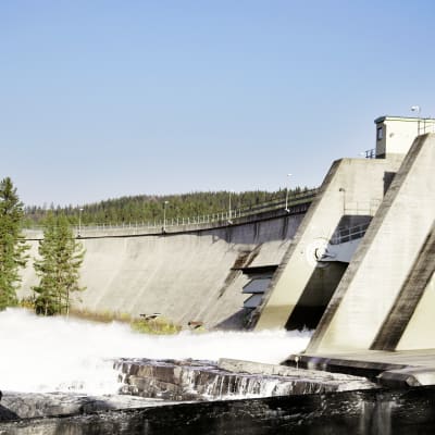 En femton meter hög damm i en fors, till vänster en skog, och till höger ett vattenkraftverk inbyggt i dammen med vatten forsande ut ur det.