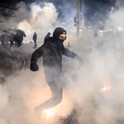 Mustaan pukeutunut ja naamioitunut mielenosoittaja kulkee kyynelkaasupilven läpi öisessä Pariisissa.