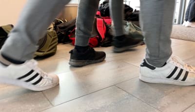 Ryggsäckar ligger på ett golv och skolelever går förbi, det syns jeansklädda ben och gymnastikskor.