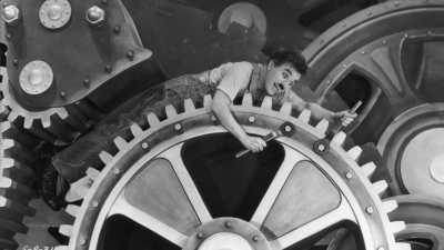 Scen från Charles Chaplins film Moderna tider, med Chaplin liggande på ett stort kugghjul.