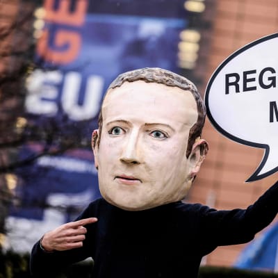 Mielenosoittaja jolla on Mark Zuckerberg naamari kasvoillaan pitäen pahvista puhekuplaa "regulate me!"