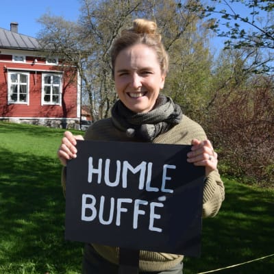 Framför ett rött hus står en leende kvinna med en skylt "Humle Buffet"