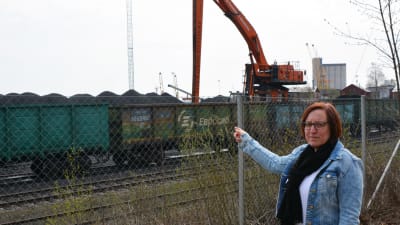 En kvinna står vid ett staket och pekar. Bakom staketet synns tågvagnar, stora kolhögar och en arbetsmaskin.