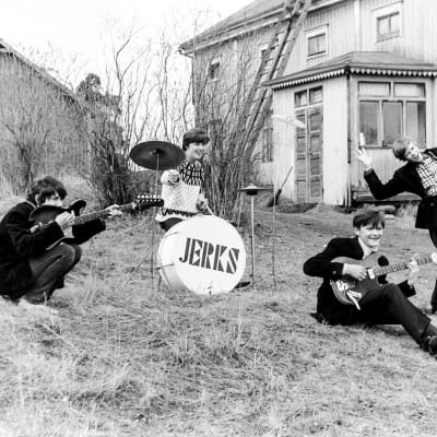 Mustavalkoisessa kuvassa nuorisoyhtye vanhab maalaistalon rinteisellä pihamaalla soittimineen. Bassorummussa lukee "Jerks"
