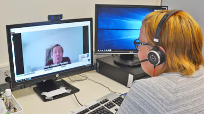 En kvinna sitter framför en dator. Hon har hörlurar på sig. På skärmen framför henne syns en äldre man som hon pratar med.