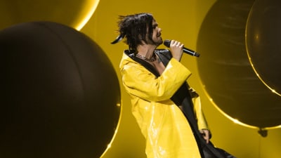En man i gul regnrock sjunger på en scen. Bakgrunden är svart.
