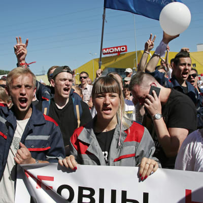 Suuri joukko työläisiä kulkee kadulla. Eturivissä olevat kantavat plakaattia, jossa on venäjänkielinen teksti isoilla kirjaimilla.