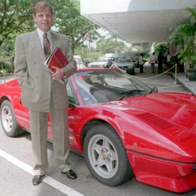 Thomas Zilliacus seisoo puku päällä punaisen henkilöauton edessä kansio kainalossaan.