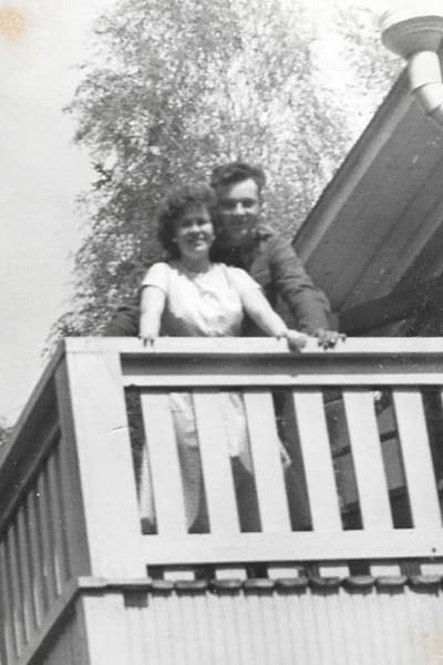 En gammal svartvit bild av en kvinna och en man som står nära varandra på en balkong och tittar in i kameran.