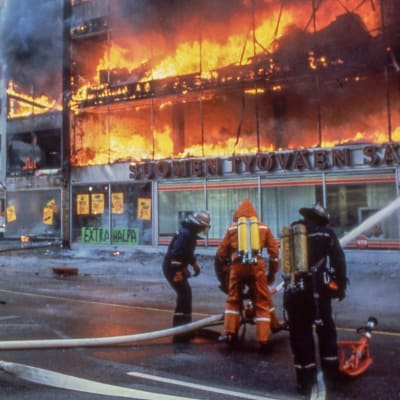 Tre brandmän, två i mörkblå kläder och en i orange kläder, står på en gata och sprutar vatten på ett höghus som är helt övertänt. På fasaden står Extra halpa och Suomen työväen säästöpankki.